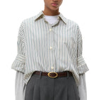 A person wears the TWP Next Ex Dancing Striped Cotton Shirt over a gray long-sleeve top, tucked into gray high-waisted pants with a brown belt.
