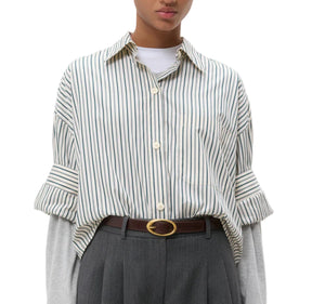 A person wears the TWP Next Ex Dancing Striped Cotton Shirt over a gray long-sleeve top, tucked into gray high-waisted pants with a brown belt.