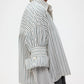 A person in the TWP Next Ex Dancing Striped Cotton Shirt with a boxy fit, layered over a grey long-sleeve top, sleeves rolled up and seen from the side.