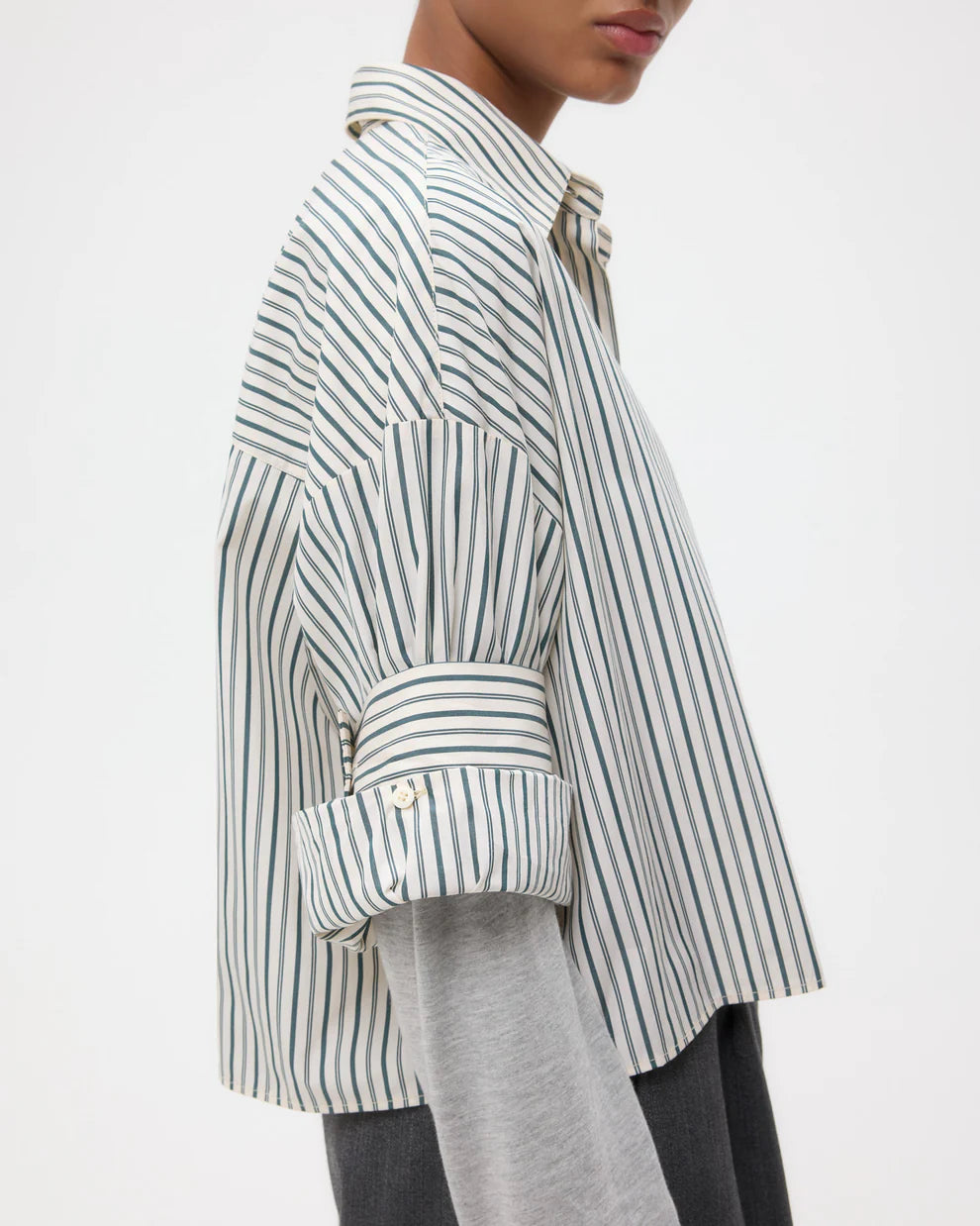 A person in the TWP Next Ex Dancing Striped Cotton Shirt with a boxy fit, layered over a grey long-sleeve top, sleeves rolled up and seen from the side.