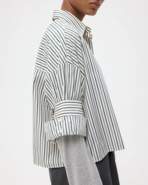 A person in the TWP Next Ex Dancing Striped Cotton Shirt with a boxy fit, layered over a grey long-sleeve top, sleeves rolled up and seen from the side.