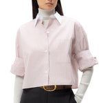 A person wearing the TWP Next Ex Classic Barre Striped Cotton Shirt—cropped, short sleeve, pink and white—over a light gray turtleneck, styled with dark trousers and a tan belt.