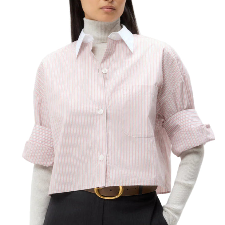 A person wearing the TWP Next Ex Classic Barre Striped Cotton Shirt—cropped, short sleeve, pink and white—over a light gray turtleneck, styled with dark trousers and a tan belt.