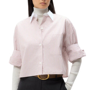A person wearing the TWP Next Ex Classic Barre Striped Cotton Shirt—cropped, short sleeve, pink and white—over a light gray turtleneck, styled with dark trousers and a tan belt.