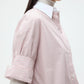 A person wears the TWP Next Ex Classic Barre Striped Cotton Shirt—a light pink and white cropped button down in fine Italian cotton—over a light grey turtleneck, sleeves rolled up to highlight its boxy silhouette.