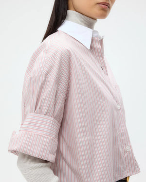 A person wears the TWP Next Ex Classic Barre Striped Cotton Shirt—a light pink and white cropped button down in fine Italian cotton—over a light grey turtleneck, sleeves rolled up to highlight its boxy silhouette.