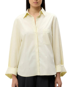 A woman wears the TWP Next Morning Shirt—an oversized light yellow Italian cotton button-down with chest pocket and wide sleeves—paired with dark pants for a relaxed look.