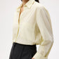 A person wearing the TWP Next Morning Shirt—an oversized, light yellow Italian cotton button-down with a front pocket—paired with black high-waisted trousers, standing against a plain background.