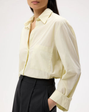 A person wearing the TWP Next Morning Shirt—an oversized, light yellow Italian cotton button-down with a front pocket—paired with black high-waisted trousers, standing against a plain background.