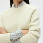 A person with dark hair wears the TWP New Jill Crewneck Sweater in cream over a collared, striped shirt, standing with arms crossed.