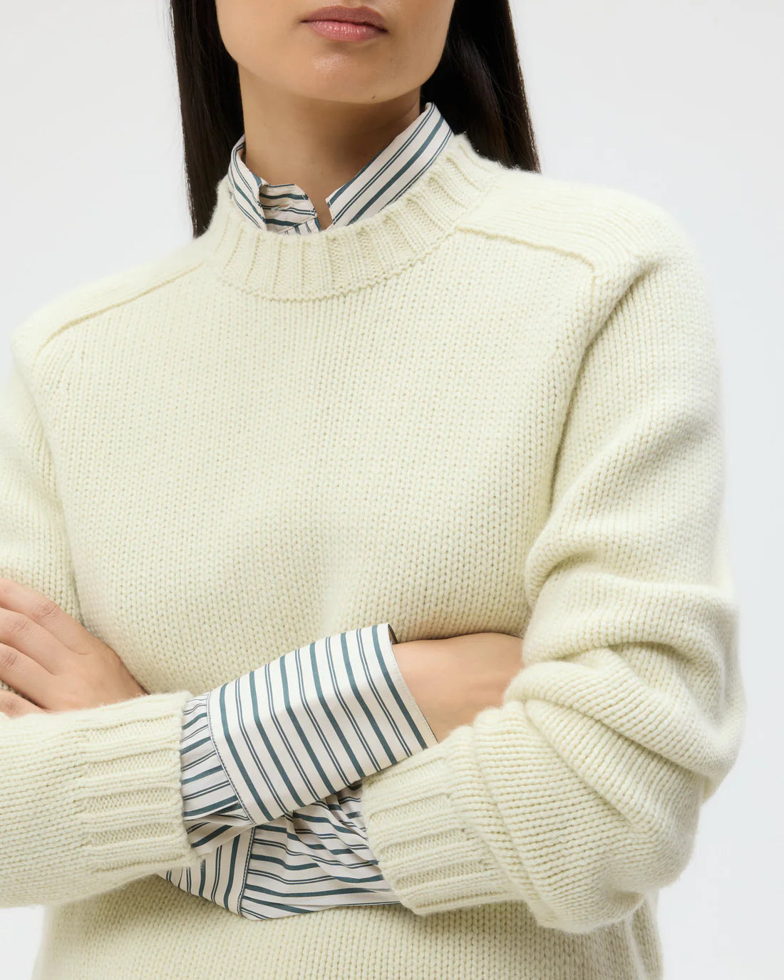 A person with dark hair wears the TWP New Jill Crewneck Sweater in cream over a collared, striped shirt, standing with arms crossed.