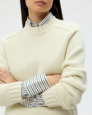 A person with dark hair wears the TWP New Jill Crewneck Sweater in cream over a collared, striped shirt, standing with arms crossed.