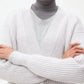 A person wears the TWP Cropped Cashmere Cardi in white, crafted by an Italian heritage mill, layered over a grey turtleneck with arms crossed, shown from shoulders to chest against a plain background.