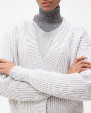 A person wears the TWP Cropped Cashmere Cardi in white, crafted by an Italian heritage mill, layered over a grey turtleneck with arms crossed, shown from shoulders to chest against a plain background.