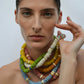 A woman with a neutral expression wears the colorful, chunky Laguna Necklace by Lizzie Fortunato, featuring bold beads, and rests her hand near her face against a plain background.