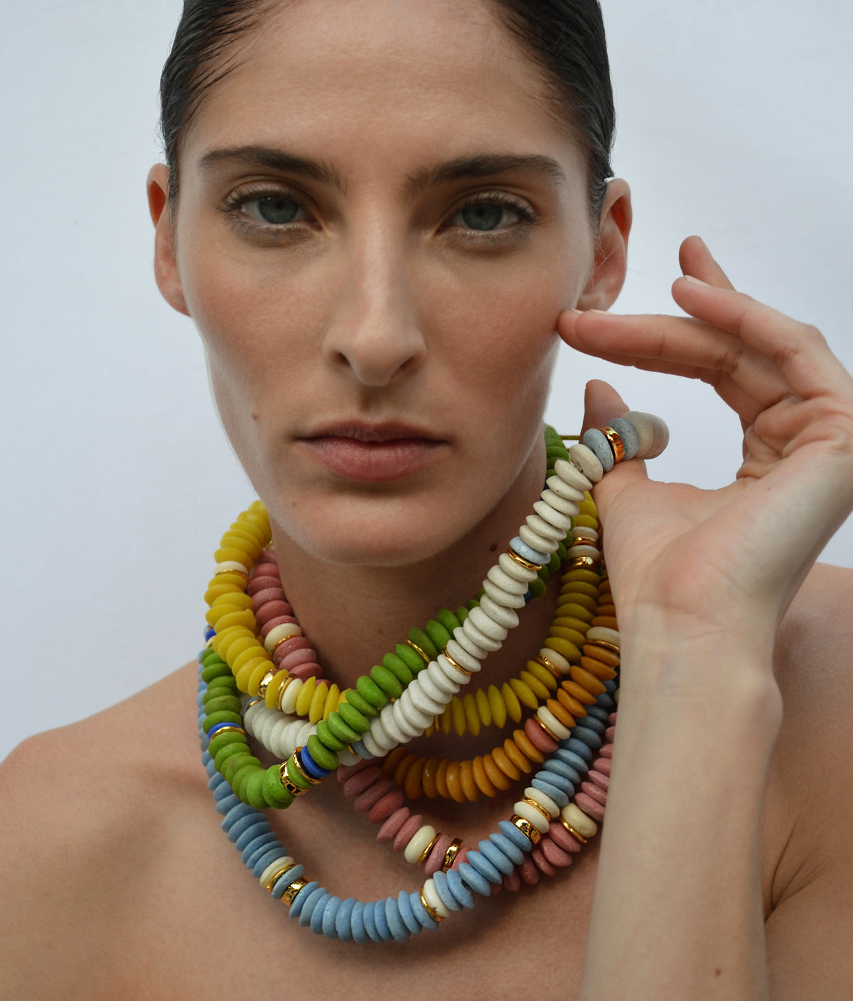 A woman with a neutral expression wears the colorful, chunky Laguna Necklace by Lizzie Fortunato, featuring bold beads, and rests her hand near her face against a plain background.