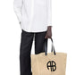 Person wearing a white button-up shirt, black wide-leg pants, and black sandals holds the Anine Bing Large Rio Tote, crafted from woven seagrass with black handles and a signature ANINE BING monogram.