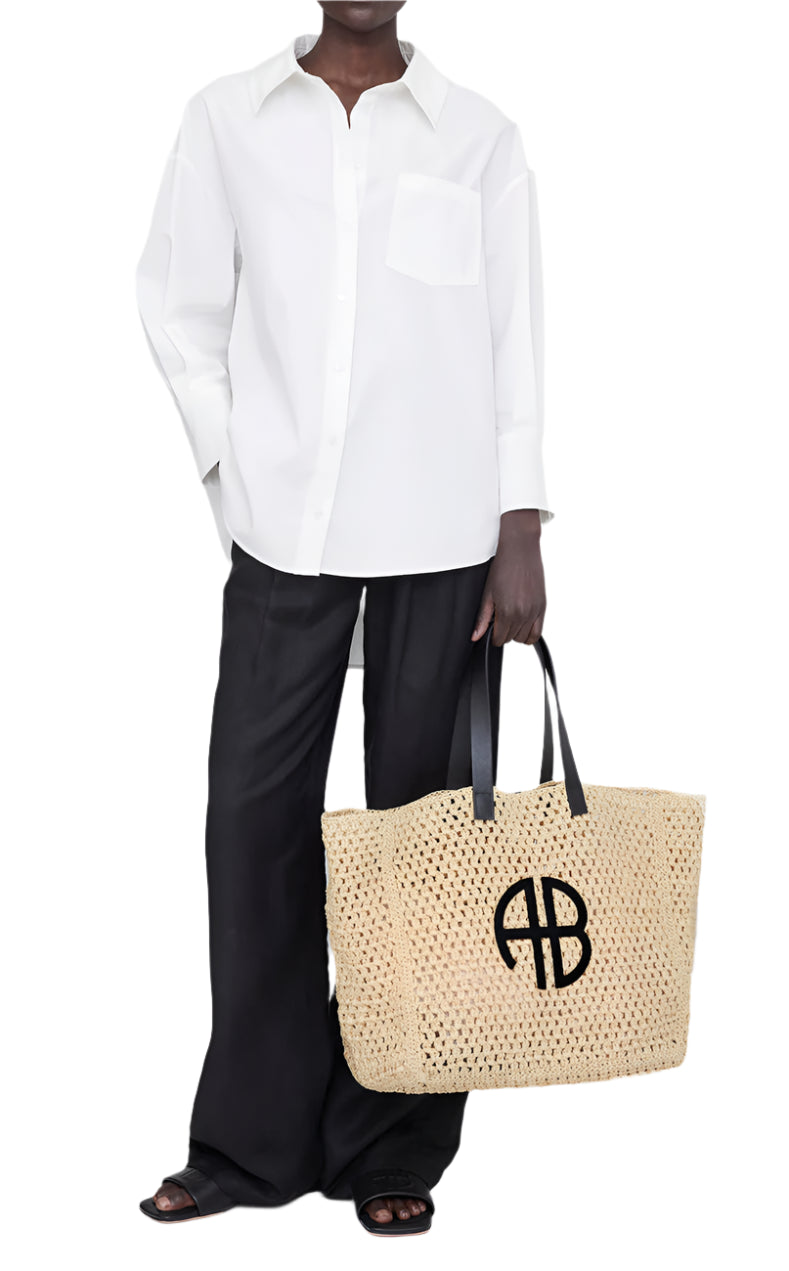 Person wearing a white button-up shirt, black wide-leg pants, and black sandals holds the Anine Bing Large Rio Tote, crafted from woven seagrass with black handles and a signature ANINE BING monogram.