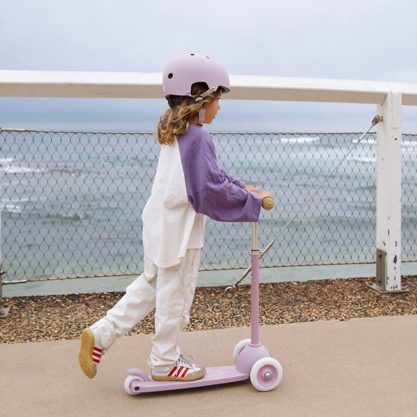 A child wearing a helmet and purple sweatshirt rides the Banwood Kids’ Eco Scooter with Basket along a paved path by the ocean, with a fence and cloudy sky in the background.
