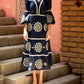 A person stands on stone steps wearing the Andres Otalora Mapa Midi Dress, a navy blue dress adorned with white floral embroidery and lantern sleeves. The background features a pink wall and a brick column.