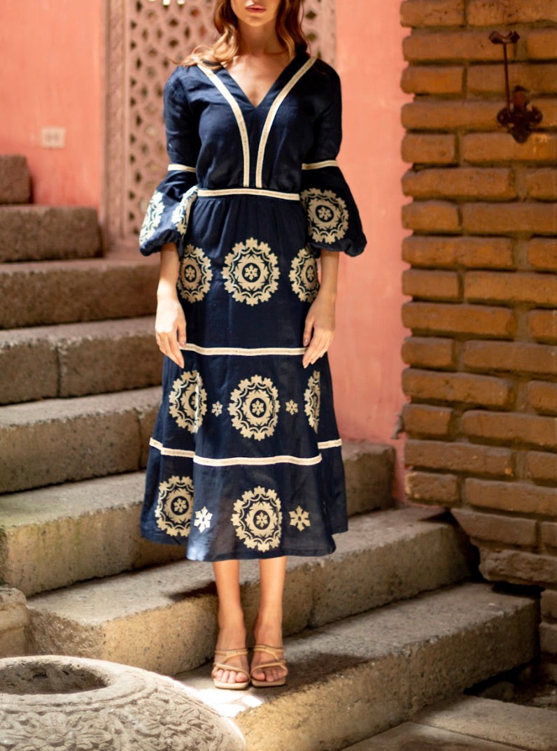 A person stands on stone steps wearing the Andres Otalora Mapa Midi Dress, a navy blue dress adorned with white floral embroidery and lantern sleeves. The background features a pink wall and a brick column.