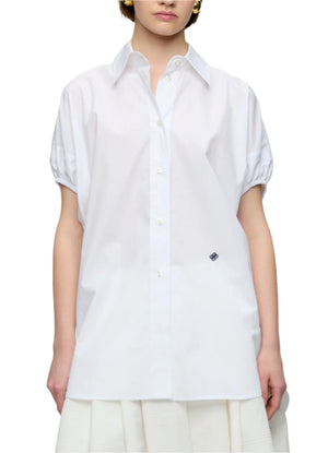 A person wearing the Callas Milano Lecce Top, a relaxed fit Italian poplin shirt with a collar and small lower-front emblem, paired with a white skirt.