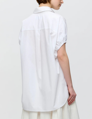 A person stands facing away, wearing the Callas Milano Lecce Top, a relaxed fit Italian poplin short-sleeve button-front shirt, paired with a white skirt against a plain background.