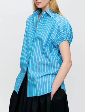 A person wearing the Callas Milano Lecce Stripe Top, a relaxed fit blue and white short-sleeve button-up shirt, paired with a black skirt.