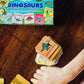 A child’s hand reaches for a stack of Stegosaurus cards beside an Eeboo Dinosaurs Memory & Matching Game box on a wooden surface.