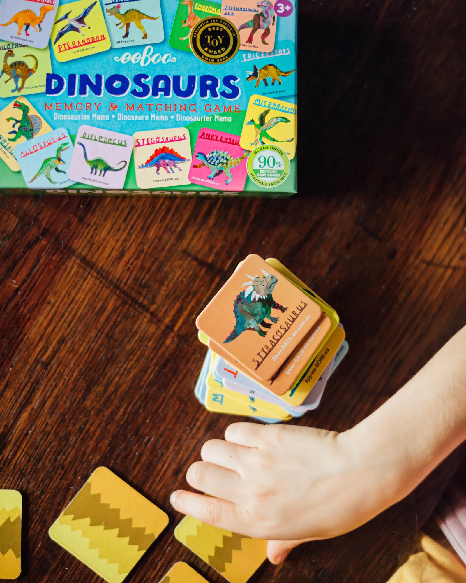 A child’s hand reaches for a stack of Stegosaurus cards beside an Eeboo Dinosaurs Memory & Matching Game box on a wooden surface.