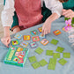 Two children play the Eeboo Garden Little Square Memory Game at a table. The game pieces and box, made with recycled board, are spread out on a teal tablecloth, with a small flower centerpiece visible in the corner.