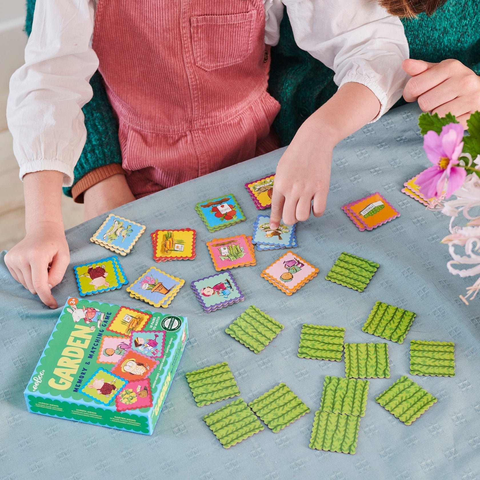 Two children play the Eeboo Garden Little Square Memory Game at a table. The game pieces and box, made with recycled board, are spread out on a teal tablecloth, with a small flower centerpiece visible in the corner.