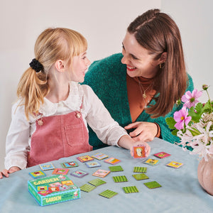 A woman and a young girl with blonde hair are seated at a table, playing the Eeboo Garden Little Square Memory Game together. The game box and cards, made with recycled board, are visible. There is a vase of flowers on the table. They are smiling at each other.