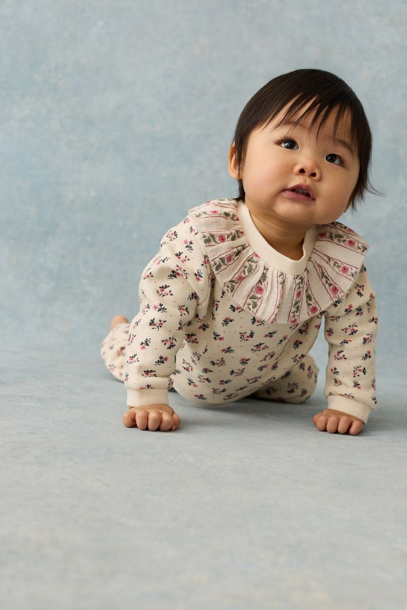A baby wearing the Louise Misha Baby Girls’ Rosaline Sweatshirt crawls on a light blue surface, gazing slightly upward with a neutral expression.