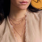 A woman wears layered gold and purple bead necklaces with small charms, including the Gigi Clozeau Clover Lumiere Diamond Pendant in 18k yellow gold, styled with a beige frayed-edge top against a yellow background.