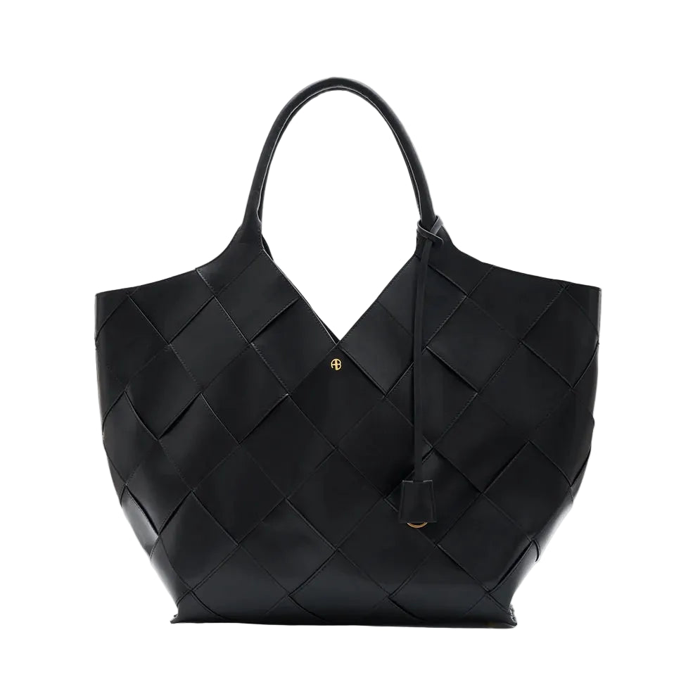 The Lili Braided Leather Tote by Anine Bing is a spacious black bag crafted from leather with a braided finish, featuring two top handles and adorned with a small gold emblem on the front, photographed against a white background.