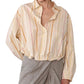A woman wearing the Lille Top by Callas Milano, a loose long-sleeve shirt with a point collar and pastel vertical stripes, paired with a light gray wrap skirt.