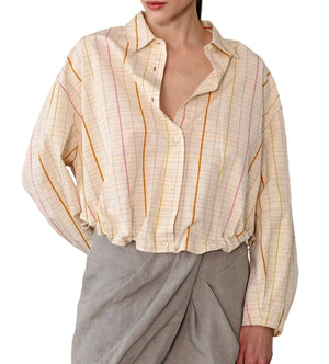 A woman wearing the Lille Top by Callas Milano, a loose long-sleeve shirt with a point collar and pastel vertical stripes, paired with a light gray wrap skirt.