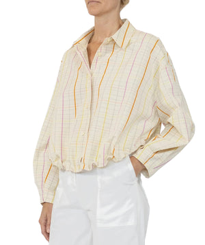 A person wearing the Callas Milano Lille Top—a loose, long-sleeve cotton poplin shirt with pastel stripes and a point collar—paired with white pants, photographed from the waist up.