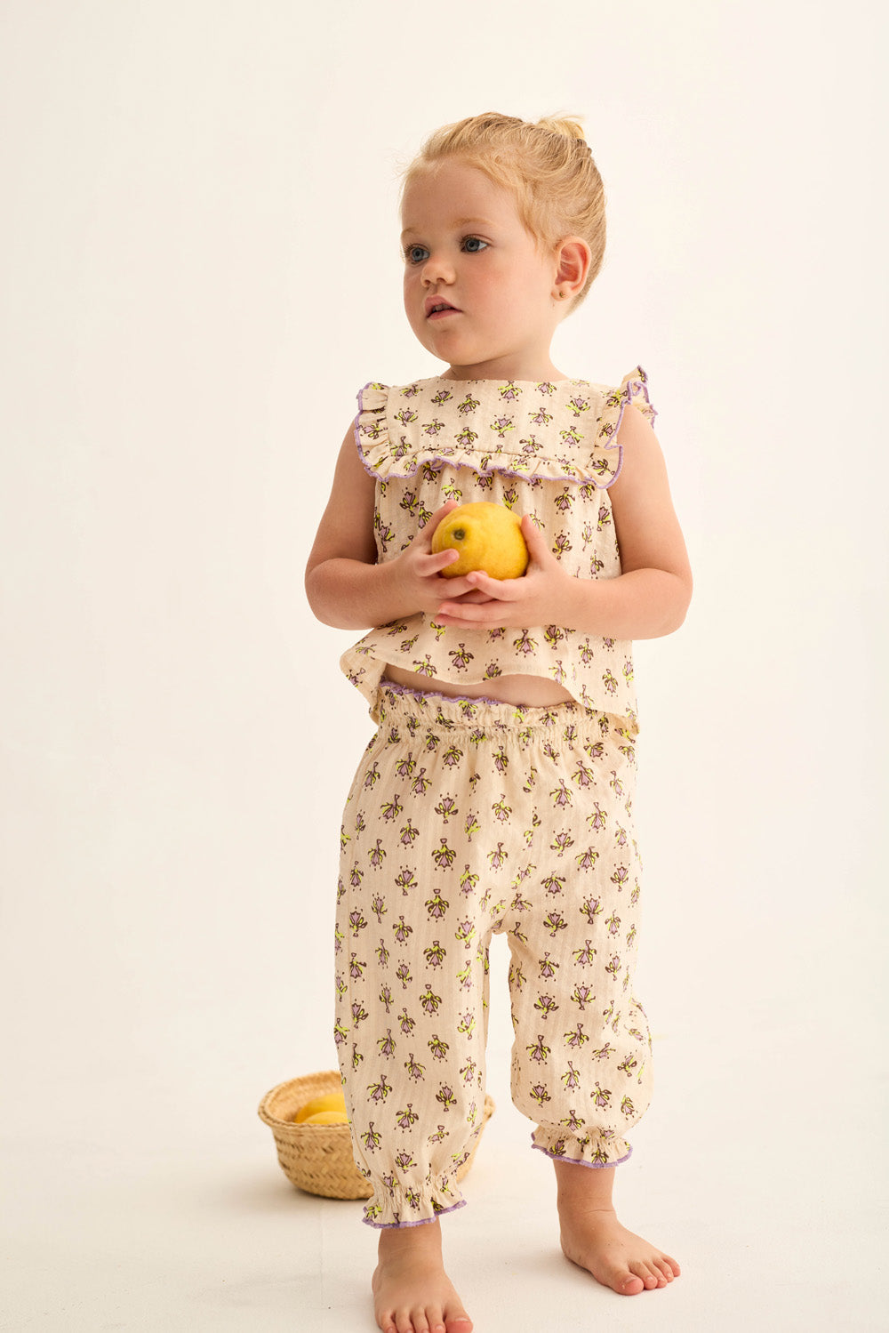 A young child stands barefoot holding a yellow apple, wearing Cozmo’s Baby Girls' Lina Pants in a cream-colored floral print with lettuce trim. Behind her on the floor is a small basket filled with more apples.
