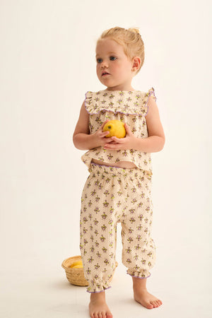 A young child stands barefoot holding a yellow apple, wearing Cozmo’s Baby Girls' Lina Pants in a cream-colored floral print with lettuce trim. Behind her on the floor is a small basket filled with more apples.