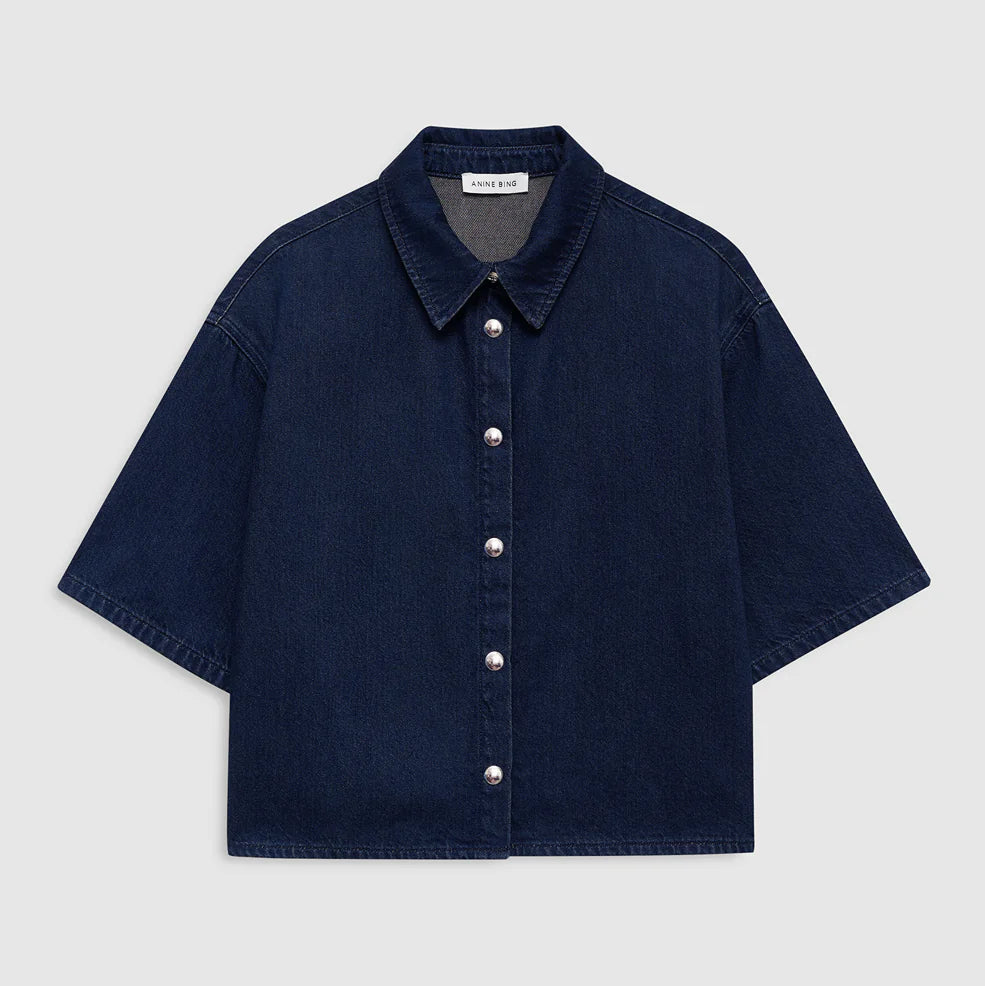 The Anine Bing Lincoln Shirt, a dark blue lightweight denim piece featuring short sleeves, a collar, and front pearl buttons, is displayed laid flat on a plain light background.