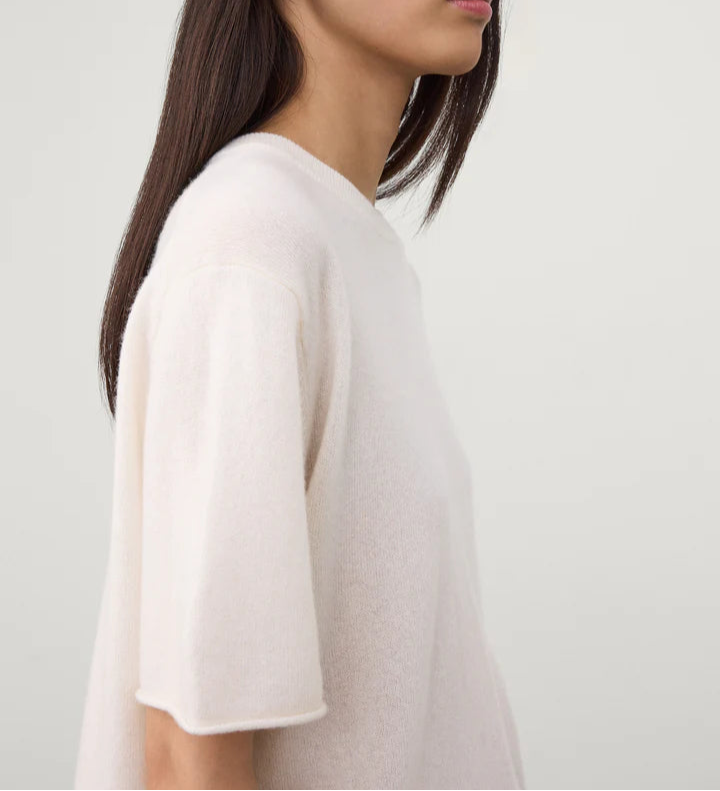 A person with straight, dark hair is shown in profile against a plain light background, wearing Lisa Yang’s Cila Cashmere T-Shirt—a loose, short-sleeved, crew-neck top in light beige.