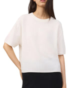 A woman wears the Lisa Yang Cila Cashmere T-Shirt—a loose-fitting, crew neck, light-colored top with short sleeves—paired with dark pants, standing with one hand on her hip.