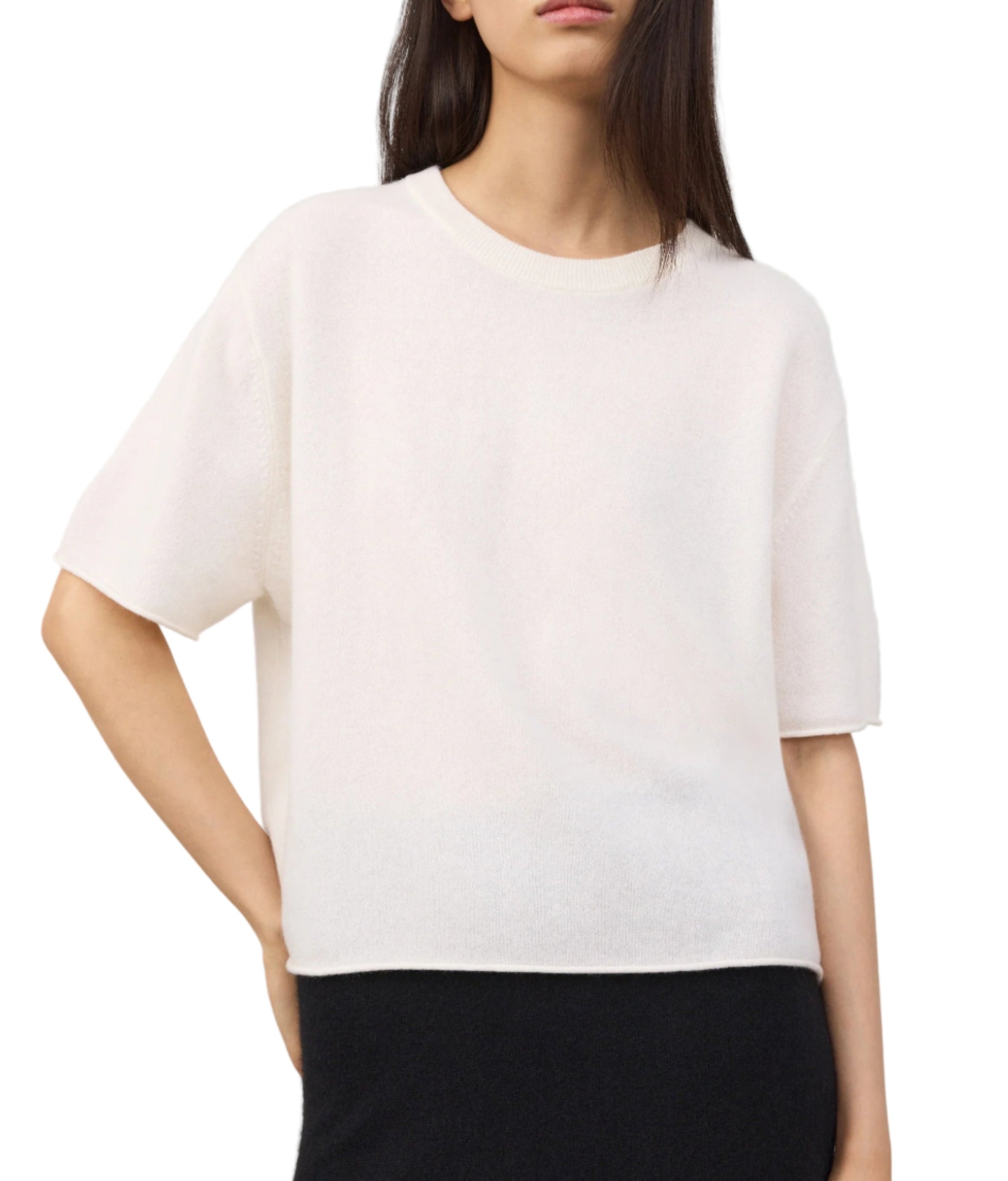 A woman wears the Lisa Yang Cila Cashmere T-Shirt—a loose-fitting, crew neck, light-colored top with short sleeves—paired with dark pants, standing with one hand on her hip.