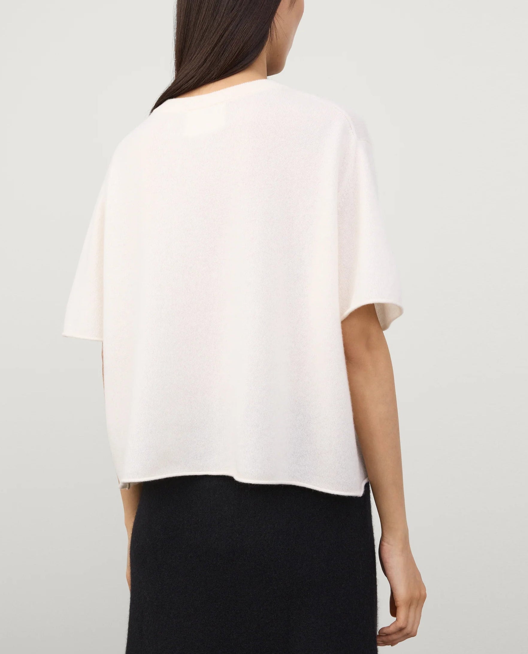 A woman with long dark hair is shown from the back, wearing the Lisa Yang Cila Cashmere T-Shirt in white and a black skirt against a plain background.