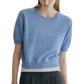 A woman is wearing the Genevra T-Shirt by Lisa Yang, a light blue, short-sleeve knit sweater featuring voluminous sleeves and a white band at the hem, paired with a black skirt—a modern take on women’s fashion.