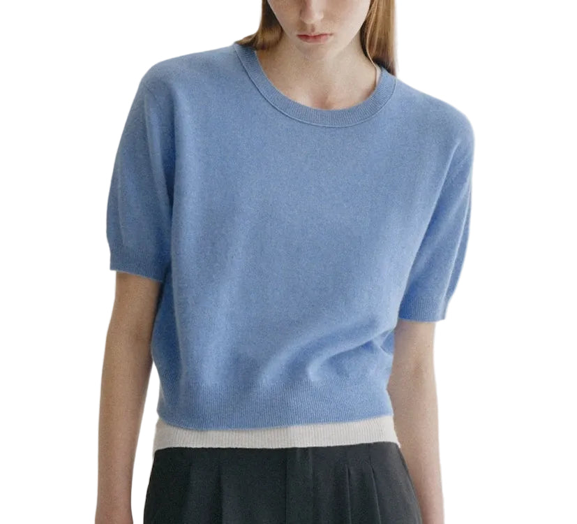 A woman is wearing the Genevra T-Shirt by Lisa Yang, a light blue, short-sleeve knit sweater featuring voluminous sleeves and a white band at the hem, paired with a black skirt—a modern take on women’s fashion.