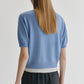 A person with straight, long light brown hair is seen from behind, wearing the Lisa Yang Genevra T-Shirt—a women’s fashion cashmere sweater in light blue with a white hem—paired with black pants, standing against a plain white background.