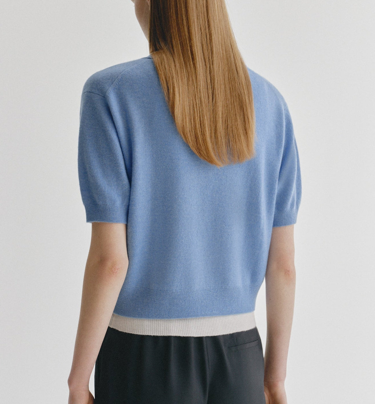 A person with straight, long light brown hair is seen from behind, wearing the Lisa Yang Genevra T-Shirt—a women’s fashion cashmere sweater in light blue with a white hem—paired with black pants, standing against a plain white background.